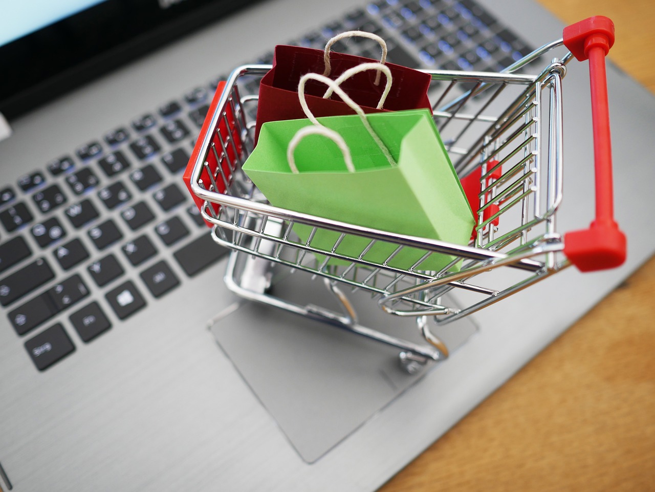 Mały wózek sklepowy z zakupami stojący na klawiaturze laptopa symbolizujący zakupy online